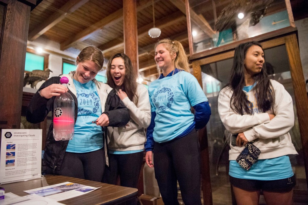 Colby College sophomore Lauren Walters, left, creates a cloud in a bottle and illuminates it with a laser as classmates Sydney Turner, left center, and Emily Hogan, right center, and Ally Corbett, right, laugh while volunteering Saturday at the L.C. Bates Museum in Hinckley as part of the Colby Cares project.