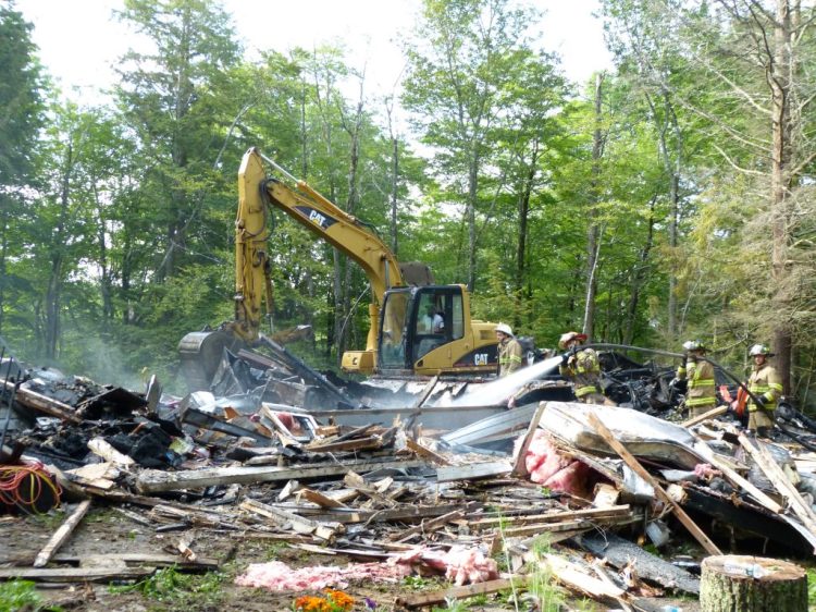An excavator digs through the remains of a Bowdoinham home destroyed by a fire the morning of June 30 as firefighters hose down smoldering debris.