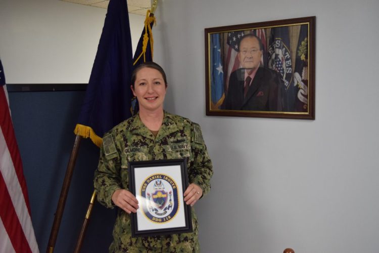 Commanding Officer DonAnn Gilmore holds the coat of arms designed for the USS Daniel Inouye while standing alongside a photo of Sen. Inouye, for whom the warship is named, at SupShip in Bath. 
