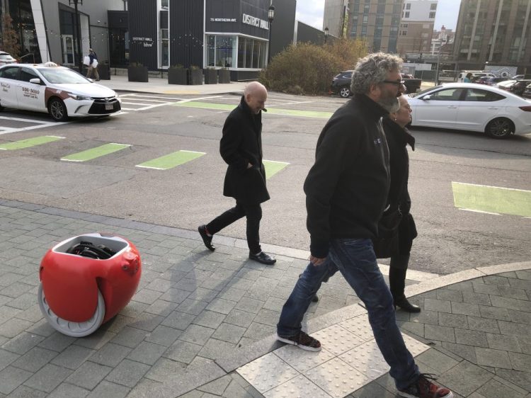 Piaggio Fast Forward CEO Greg Lynn, center, is followed by his company's Gita carrier robot as he crosses a street on Monday in Boston. The two-wheeled machine is carrying a backpack and uses cameras and sensors to track its owner. Associated Press/Matt O'Brien