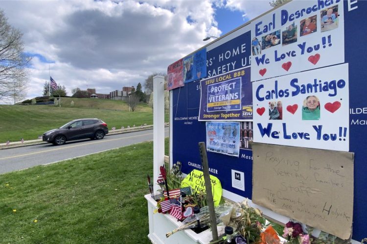 Tributes to veterans cover a sign Tuesday, near an entrance road to the Holyoke Soldiers' Home in Holyoke, Mass., where nearly 70 people have died due to the coronavirus.