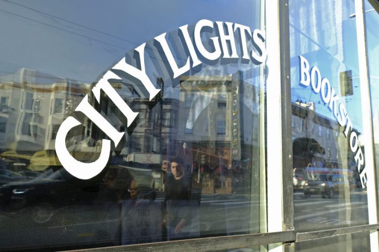 In this March 13, 2020 file photo, people are reflected in the window of the City Lights Bookstore in North Beach as they walk along Columbus Avenue in San Francisco. 