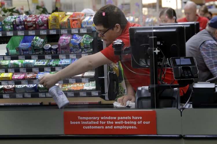 Garrett Ward sprays disinfectant on a conveyor belt between checking out shoppers behind a plexiglass panel March 26 at a Hy-Vee grocery store in Overland Park, Kan. 