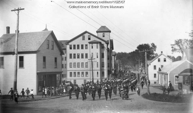 A parade crosses the bridge that spanned the Mousam River going south on Main Street in Kennebunk in this circa 1900 photo. 
The Davis Shoe Company stands as the tallest building to the left. On May 3, 1903 there was a large fire in the Davis Shoe Company building which destroyed most of the buildings in this photograph, with others nearby suffering heavy damage.