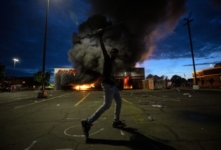 A man poses for a photo in the parking lot of an AutoZone store in flames, while protesters hold a rally for George Floyd in Minneapolis on Wednesday. Violent protests over the death of the black man in police custody broke out in Minneapolis for a second straight night.