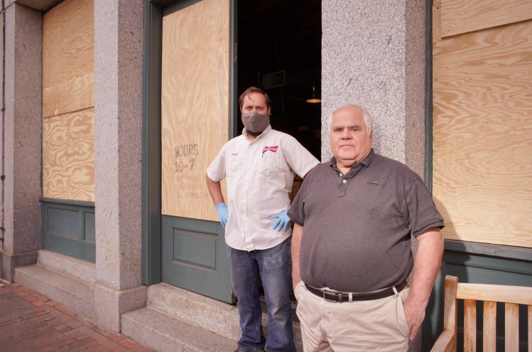 Owner Jacques deVIllier, right, and manager Kevin Casey stand in front of the boarded-up windows of Old Port Spirits and Cigars on Commercial Street in Portland on Thursday. The windows were broken Monday night by looters who stole beer and liquor from the store.