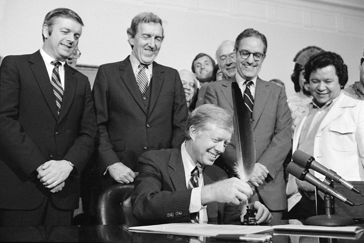 President Jimmy Carter uses an Indian quill pen to sign  the Maine Indian Claims Settlement Act of 1980 at a ceremony at the White House on Oct. 10, 1980.  From left to right are: Maine Gov. Joseph Brennan, Secretary of State Edmund Muskie, Secretary of the Interior Cecil Andrus, Maine Sen. George Mitchell, and Terrance Polchies, a leader of the Association of Aroostook Indians. 