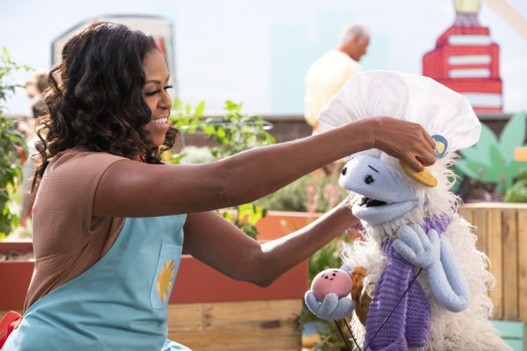 Former first lady Michelle Obama puts a chef's hat on Waffles, a furry puppet with waffle ears and holding Mochi, a pink round puppet, on the set of the children's series "Waffles + Mochi." Obama is launching the new Netflix children’s food show Tuesday.