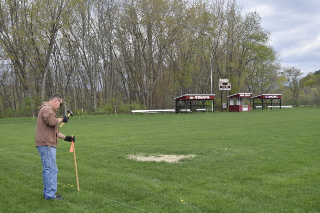 In the foreground, a man in a brown sweatshirt and blue jeans pounds a mallet into a wooden pole. He stands on a grassy field with a patch covered in tan hay. In the background, there is an electronic scoreboard and two shelters that have "University of Maine Farmington" logos on them.