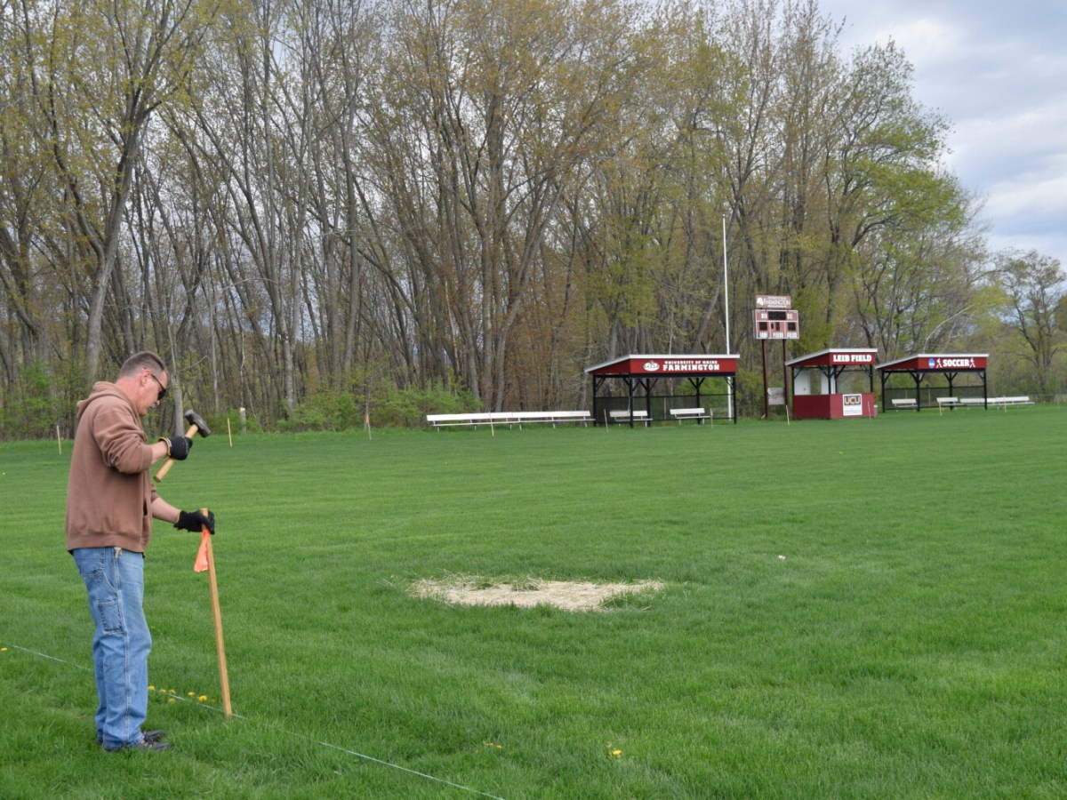 In the foreground, a man in a brown sweatshirt and blue jeans pounds a mallet into a wooden pole. He stands on a grassy field with a patch covered in tan hay. In the background, there is an electronic scoreboard and two shelters that have "University of Maine Farmington" logos on them.