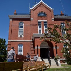Movers bring in a new cherry-wood stained bench into the Franklin County Superior Court, a red-brick building with white embellishments and big windows. In front of the movers sit benches that are being removed. The old benches are old, worn down, and fading. Behind the courthouse is a bright blue sky without any clouds.