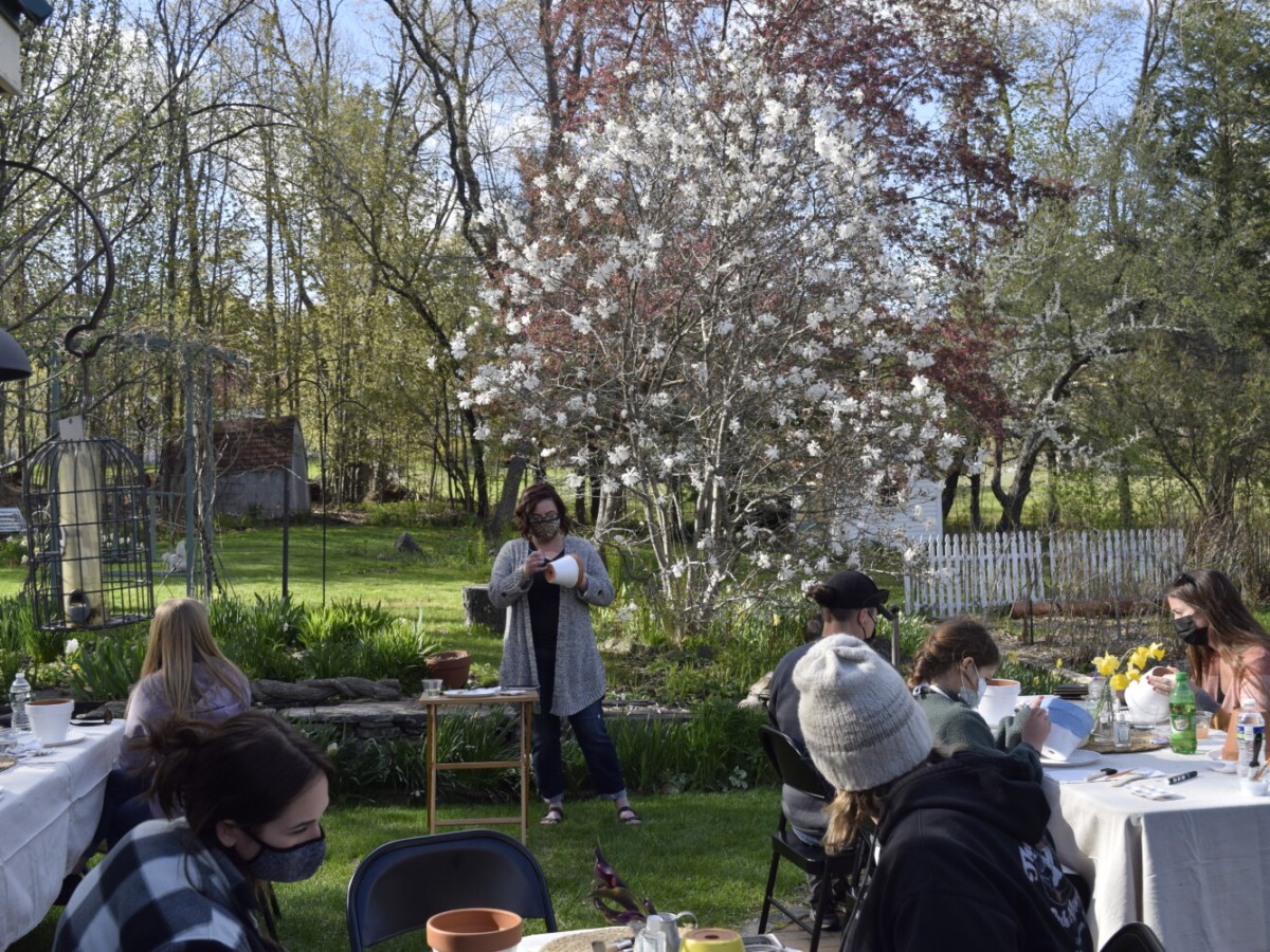 Five adults and two children sit, in a garden, at tables covered in art supplies and flower pots while a woman stands in the background painting a flower pot.