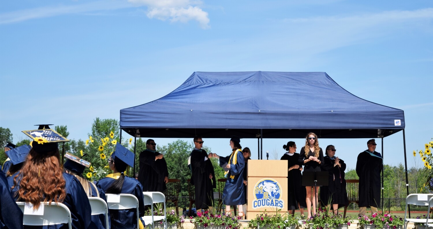 In the foreground, high school graduates sit facing a stage wearing decorated graduation caps on their heads. In the background, a student walks across a stage in a blue graduation robe and cap holding a diploma. The student is fist bumping a man in a black robe wearing sunglasses. He is surrounded by other school administrators in black robes who are clapping. The stage has a podium on it with the Mt. Blue "cougars" logo on it.