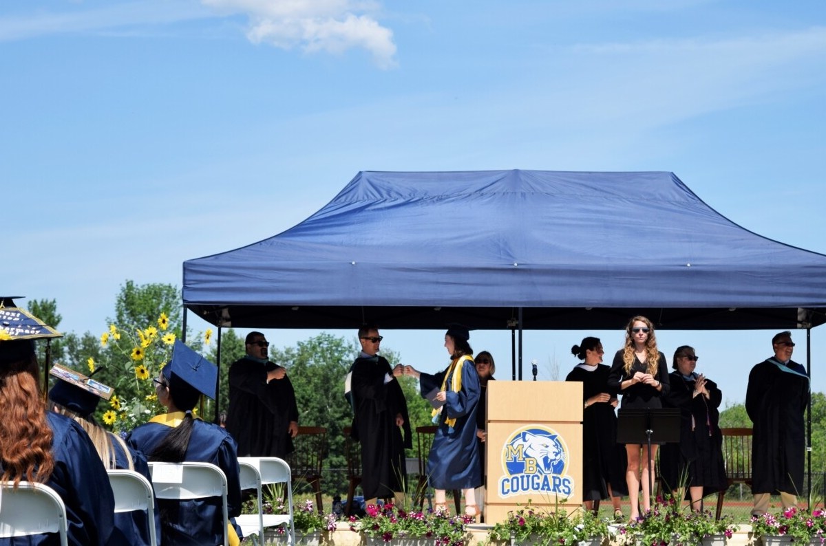 In the foreground, high school graduates sit facing a stage wearing decorated graduation caps on their heads. In the background, a student walks across a stage in a blue graduation robe and cap holding a diploma. The student is fist bumping a man in a black robe wearing sunglasses. He is surrounded by other school administrators in black robes who are clapping. The stage has a podium on it with the Mt. Blue "cougars" logo on it.