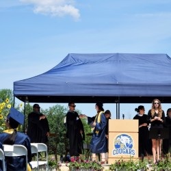 In the foreground, high school graduates sit facing a stage wearing decorated graduation caps on their heads. In the background, a student walks across a stage in a blue graduation robe and cap holding a diploma. The student is fist bumping a man in a black robe wearing sunglasses. He is surrounded by other school administrators in black robes who are clapping. The stage has a podium on it with the Mt. Blue "cougars" logo on it.