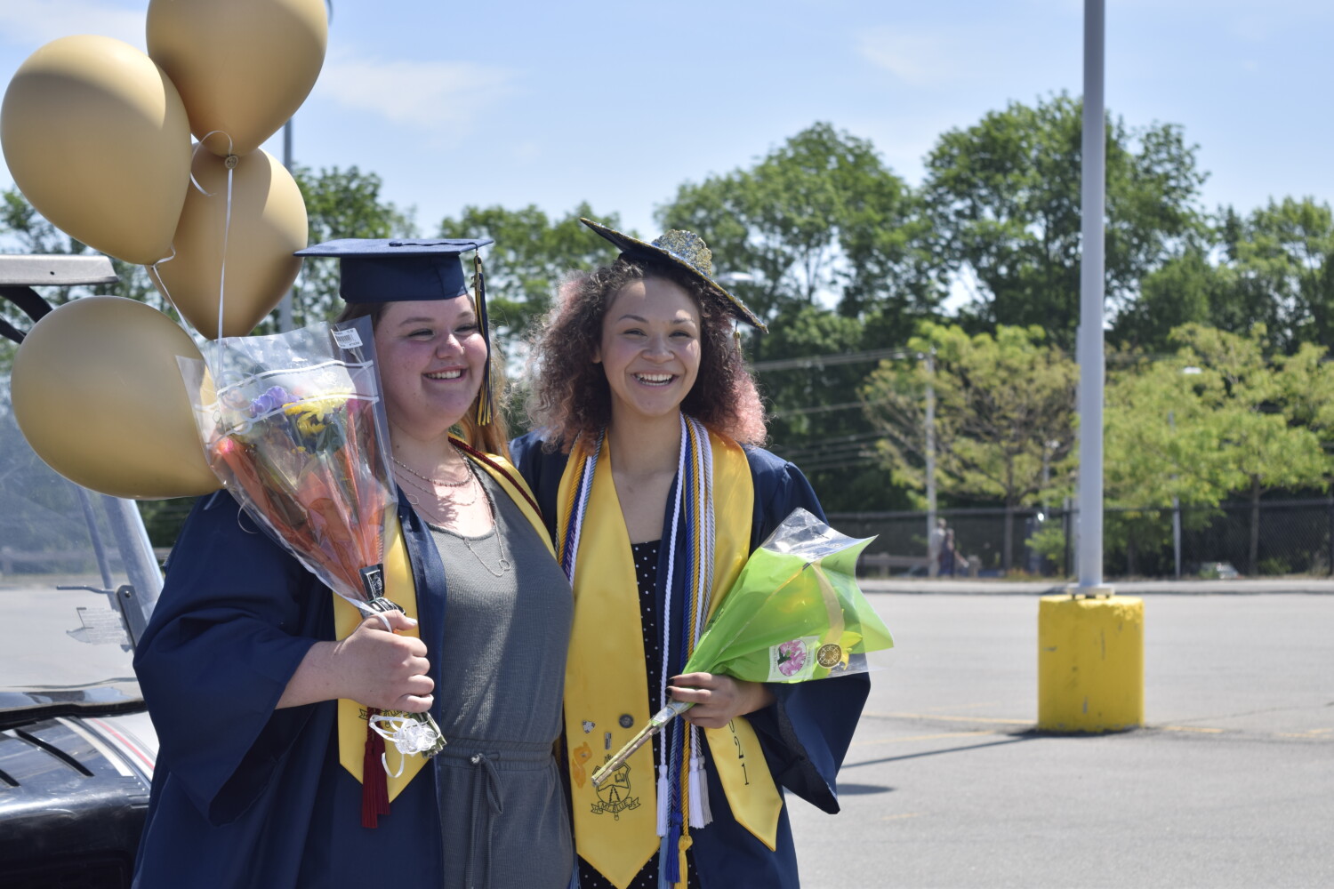 Two girls are smiling widely posing for a photo. one is wearing academic cords with a graduation robe and cap that has a crown glued on it. She is holding flowers. The girl to her right is wearing a plain graduation cap and holding gold balloons.