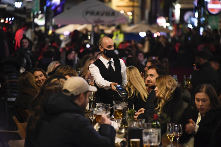 Awaiter wears a face mask as people eat and drink outside restaurants in Soho, in London, on Nov. 4, 2020. British Prime Minister Boris Johnson is expected to confirm Monday June 14, 2021, that the next planned relaxation of coronavirus restrictions in England will be delayed as a result of the spread of the delta variant first identified in India. 