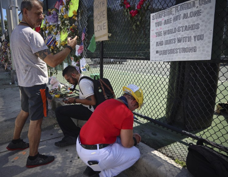 Hialeah resident Alison Kairuz, right, bows her head in prayer after pinning her hand-made sign to the fence in support of families and friends who lost love ones at the memorial site for victims of the partially collapsed South Florida condo building Champlain Towers South, in Surfside, Fla., on Sunday.
