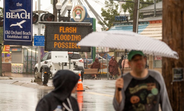 Umbrella-toting visitors walk past a sign warning of potential flash flooding as the wind and rain of Tropical storm Henri arrives on the Maine coast in Old Orchard Beach.