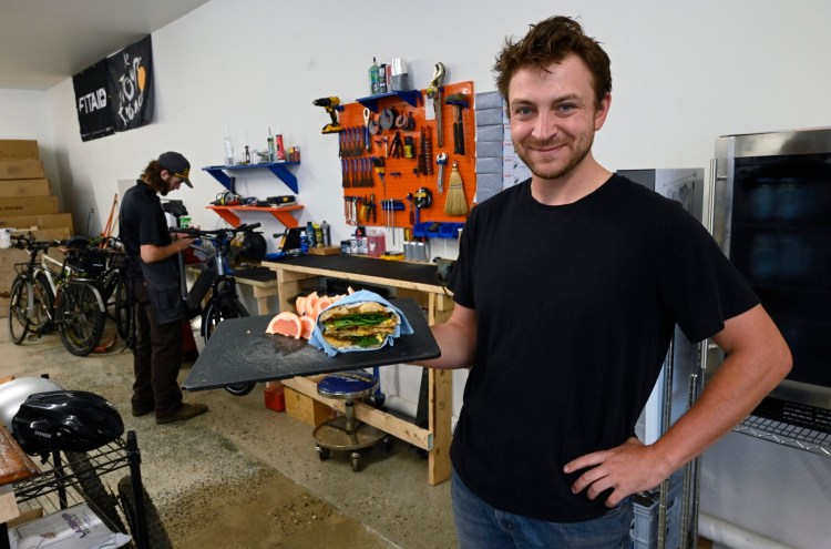 Thaddeus St. John, president of 93 Main Electric Bike Cafe, shows off a breakfast sandwich with bacon, egg, spinach and cheese on a bagel as bike mechanic Eli Falatko works on an electric bike in the shop at Electric Bike Cafe.