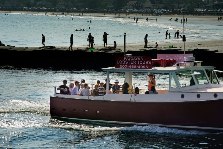 The Rugosa, a boat that provides lobster tours out of Kennebunkport, returns up the Kennebunk River after a tour on Aug. 11 while people walk and swim at Gooch’s Beach in Kennebunk on the other side of the river. With many people not taking vacations abroad, Maine has seen an increase in summer tourism.