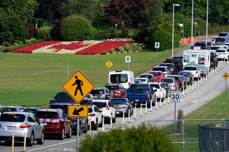 A line of vehicles wait to enter Canada at the Peace Arch border crossing in view of a Canadian flag made of flowers Monday, Aug. 9, 2021, in Blaine, Wash. Canada lifted its prohibition on Americans crossing the border to shop, vacation or visit, but America kept similar restrictions in place, part of a bumpy return to normalcy from coronavirus travel bans. (AP Photo/Elaine Thompson)
