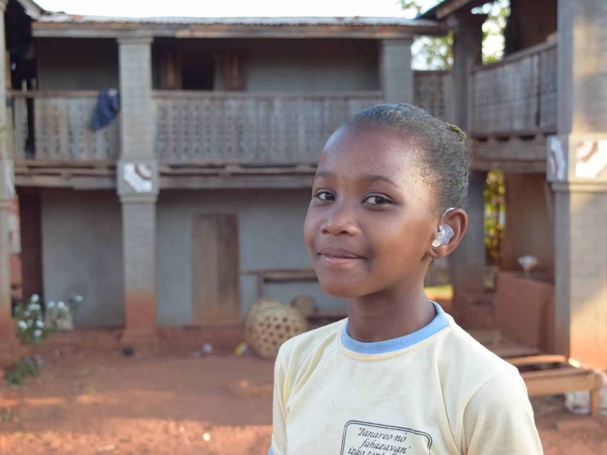 Aro is a tall 10-year-old Malagasy girl. She is wearing a yellow t-shirt from her church and her new hearing aids. She is standing in front of her home, which is three two-story brick homes connected by a shared balcony on the second floor.